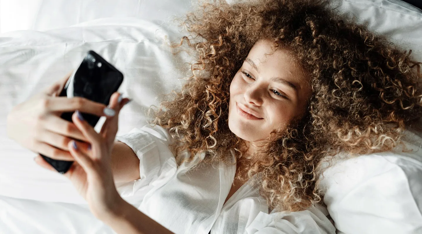 A Kabannas guest lying in bed under white sheets, smiling at her phone.